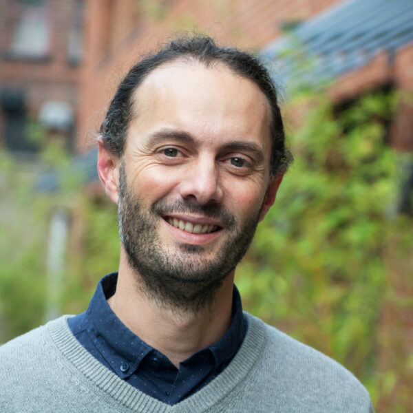 A man with a beard, dressed in a grey sweater, smiling warmly at the camera.