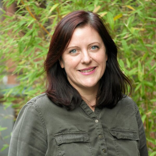 A woman with dark hair wearing a green shirt, smiling and standing against a neutral background.
